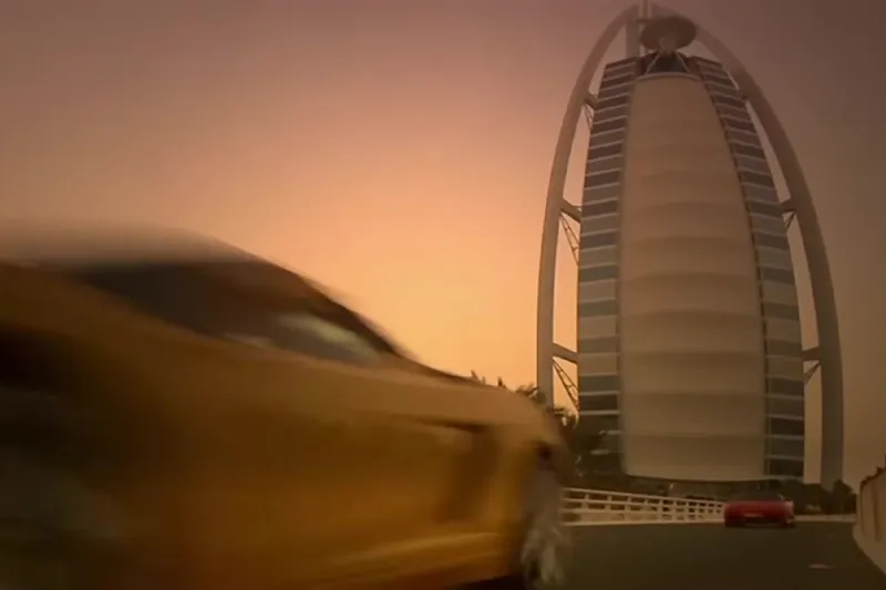 Отель Burj Al Arab на закате и желтый спортивный автомобиль, проезжающий по подъездному мосту на переднем плане.