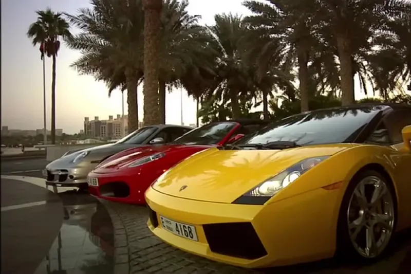 Желтый Lamborghini и красный Ferrari припаркованы перед отелем Burj Al Arab на фоне пальм и вечернего неба.