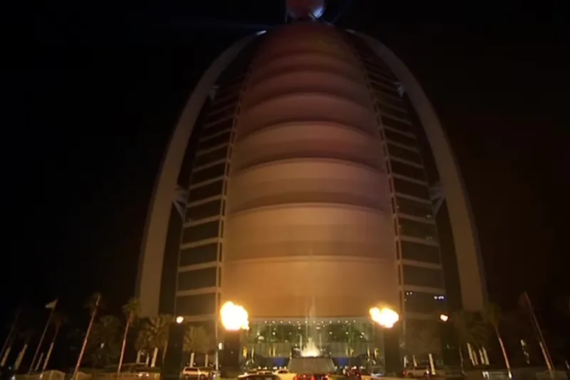 Ночной вид снизу на фасад здания Burj Al Arab, освещенный теплым светом золотых оттенков.