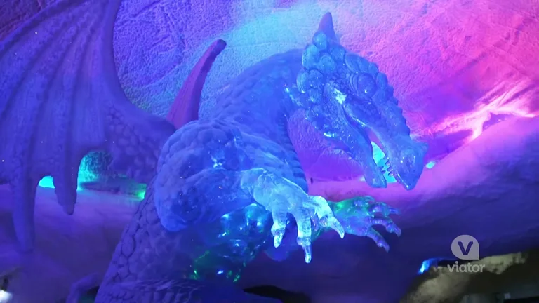 Детализированная ледяная скульптура дракона с внутренней синей и пурпурной подсветкой, расположенная в пешеходной зоне зимнего парка.