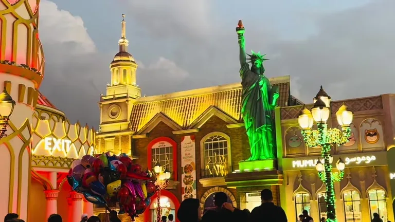 Копия статуи Свободы с зеленой подсветкой на фоне вечерних павильонов и воздушных шаров.