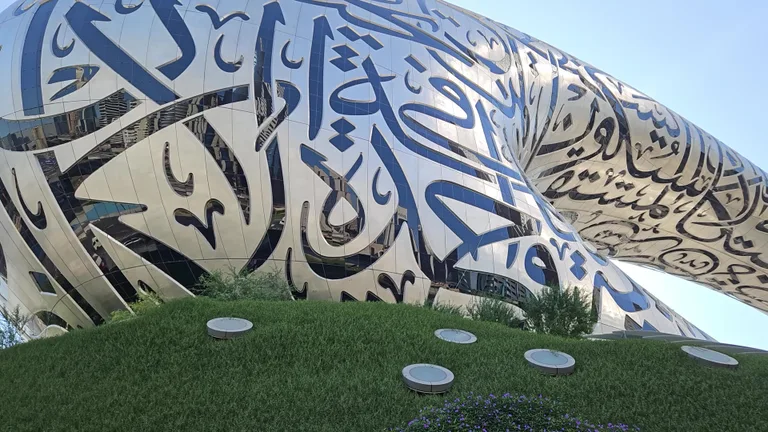 Фрагмент стального фасада с каллиграфией, возвышающегося над зеленым искусственным холмом у основания здания.