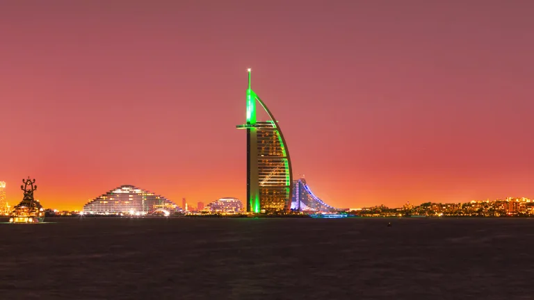 Вечерний вид на Бурдж-эль-Араб с зеленой подсветкой на фоне оранжево-пурпурного неба во время заката.