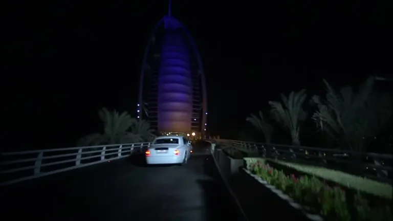 Белый автомобиль представительского класса подъезжает к зданию Бурдж-эль-Араб, освещенному фиолетовой подсветкой в ночное время.