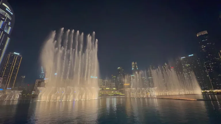 Вечернее представление подсвеченных музыкальных фонтанов на фоне современных небоскребов в центре Дубая.
