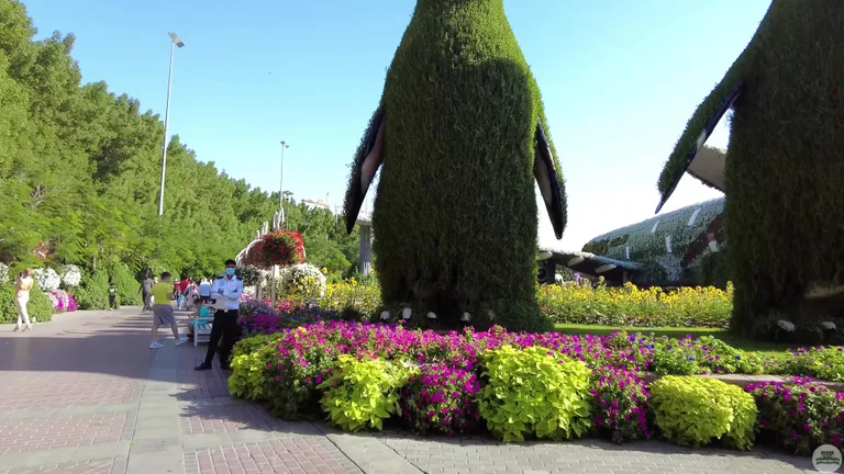 Массивные топиарные фигуры пингвинов, установленные среди поля желтых и фиолетовых цветов.