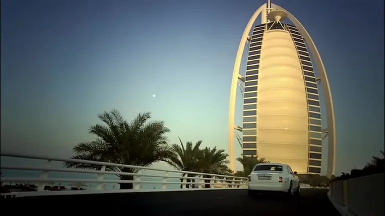 Белый автомобиль подъезжает к освещенному зданию отеля Burj Al Arab в сумерках по мосту с пальмами.