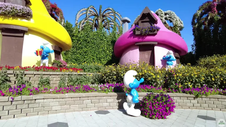 Яркие дома в форме грибов и фигуры персонажей мультфильма в окружении фиолетовых цветочных клумб.