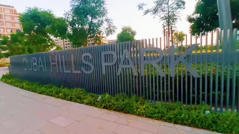 Информационная вывеска Dubai Hills Park, закрепленная на темном реечном заборе среди невысоких зеленых кустарников.