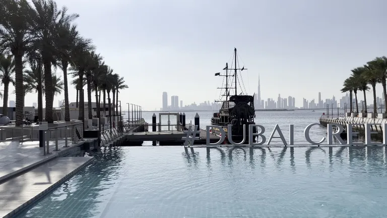 Инфинити-бассейн с надписью #DUBAICREEK на фоне традиционной лодки доу и панорамы города с башней Бурдж-Халифа.