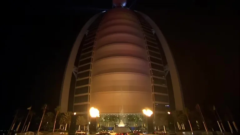 Ночной вид снизу вверх на изогнутый фасад Бурдж-эль-Араб с теплым архитектурным освещением и главным входом.