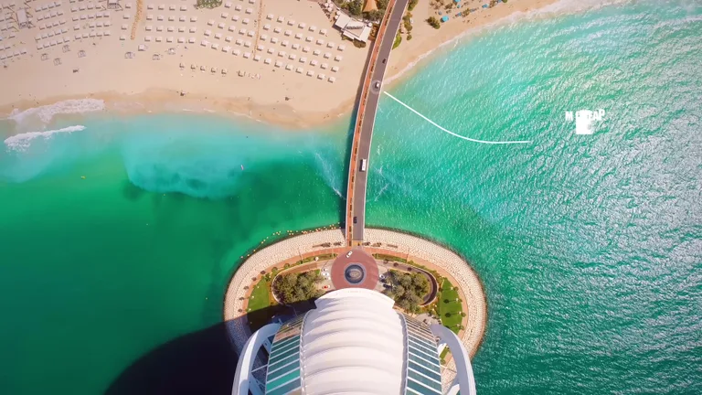 Вид сверху на Бурдж-эль-Араб, соединительный мост и бирюзовые воды залива, контрастирующие с белым песком.