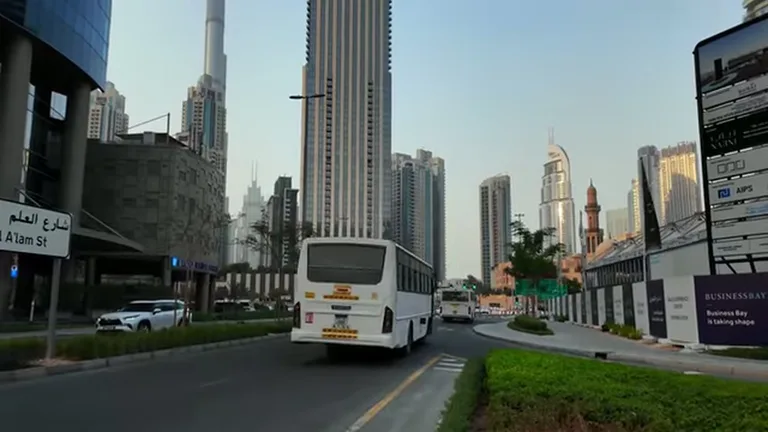 Белый автобус на изогнутой дороге в районе Business Bay на фоне современных высотных зданий и плотной городской застройки.